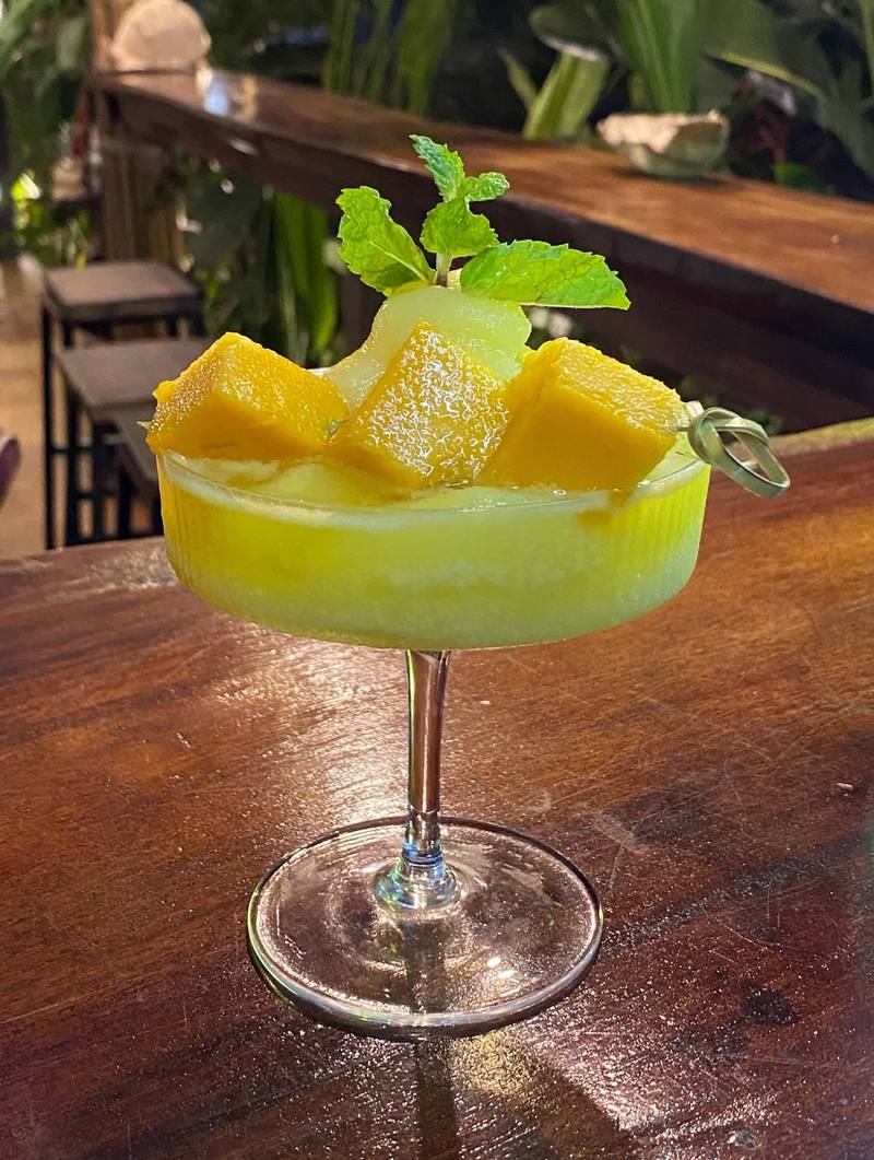 A mango dessert drink in a coupe glass with a sprig of mint on a wooden bar.