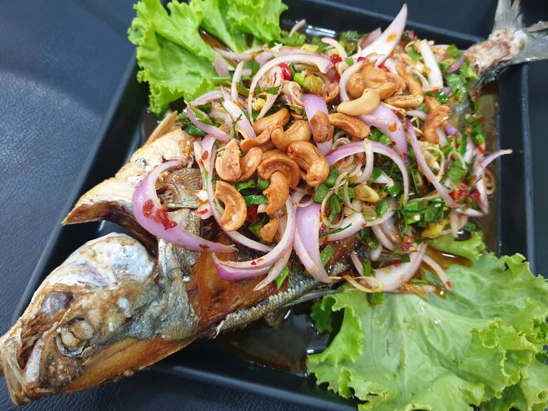 A whole grilled fish topped with a salad of red onion, cashews and herbs, on a dark plate with lettuce.